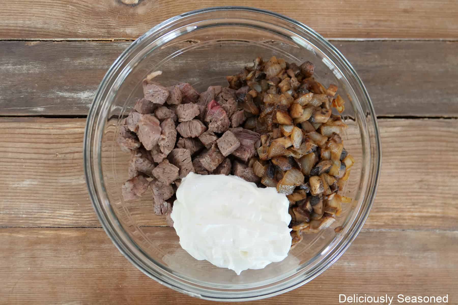 A glass bowl with cooked ribeye, sauteed onions, and sour cream in the bowl before being mixed together.