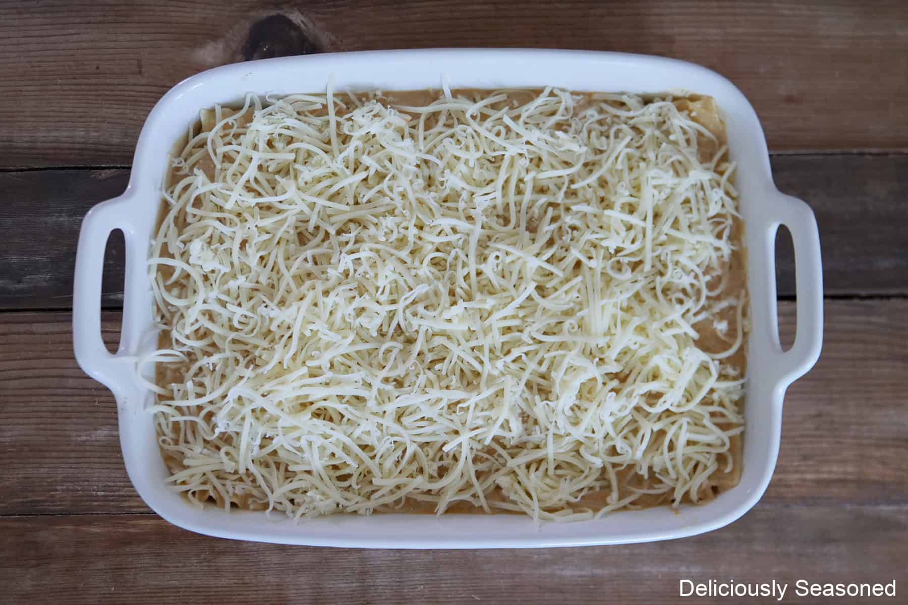 A wood surface with a white oblong baking dish filled with layers of noodles, meat, cheese, and then topped with shredded mozzarella.
