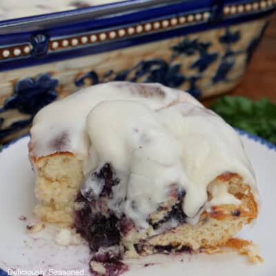 A white round plate with blue trim with a blueberry sweet roll on it with a bite taken out of it.