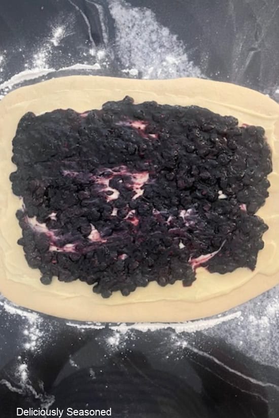 A black granite countertop with the homemade dough rolled out and covered with the cream cheese mixture and blueberry filling.