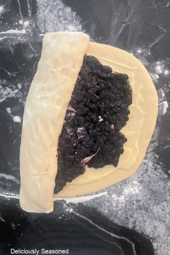 A black granite countertop with the blueberry sweet roll dough being rolled up to make the rolls.