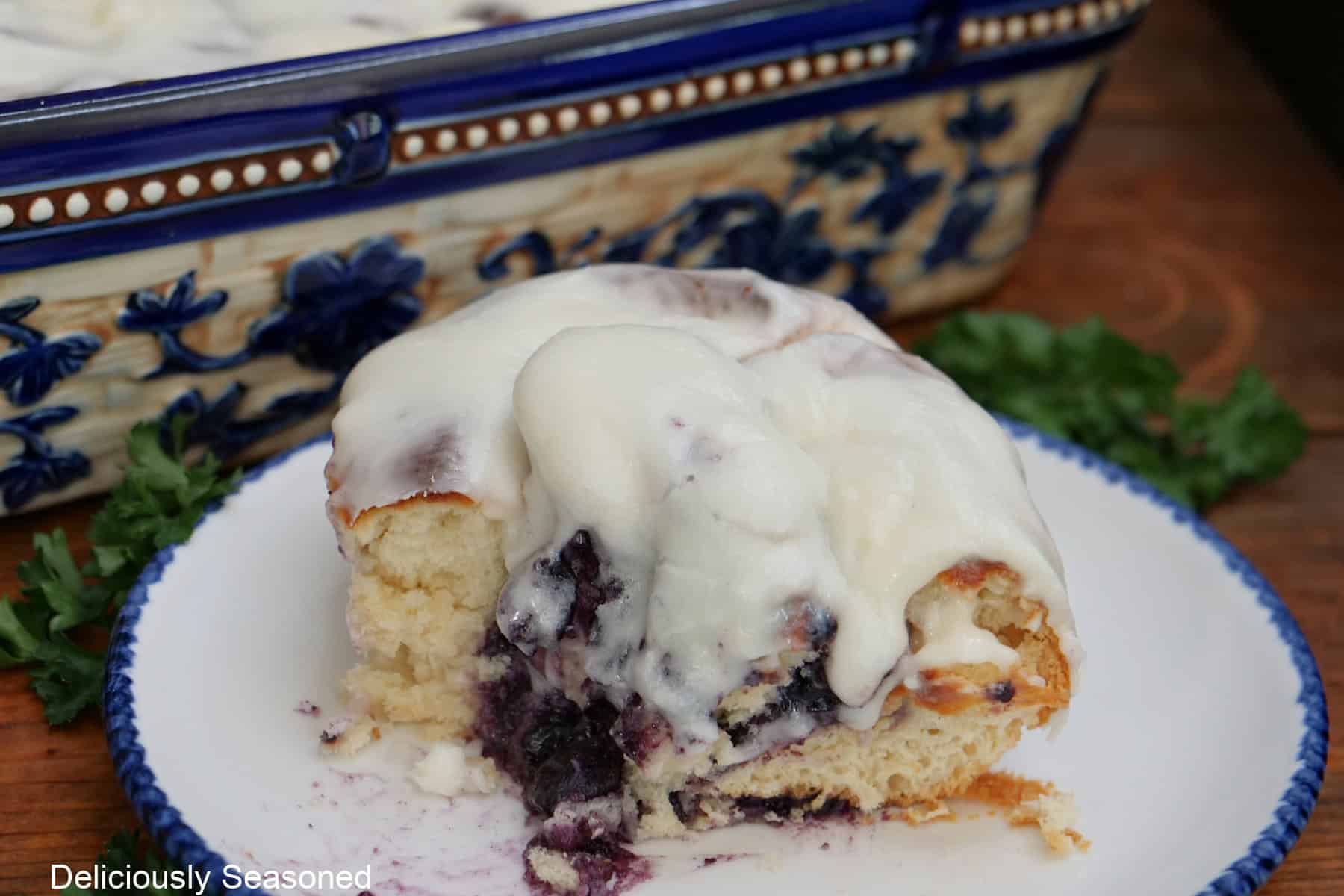 A round white plate with blue trim with a blueberry sweet roll on it with a bite taken out of it.