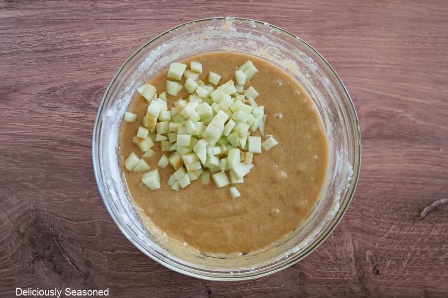 A wood surface with a glass bowl filled with blondie batter and diced apples added.