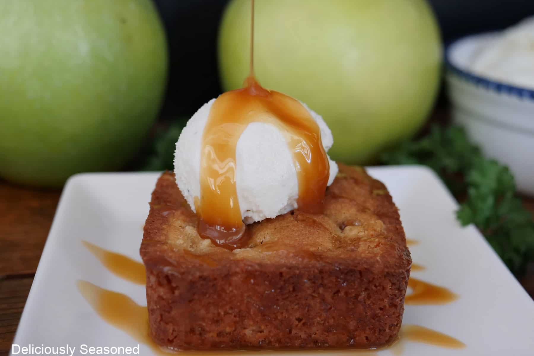 A white plate with a caramel apple blondie on it with a scoop of vanilla ice cream and caramel sauce drizzled over the top.