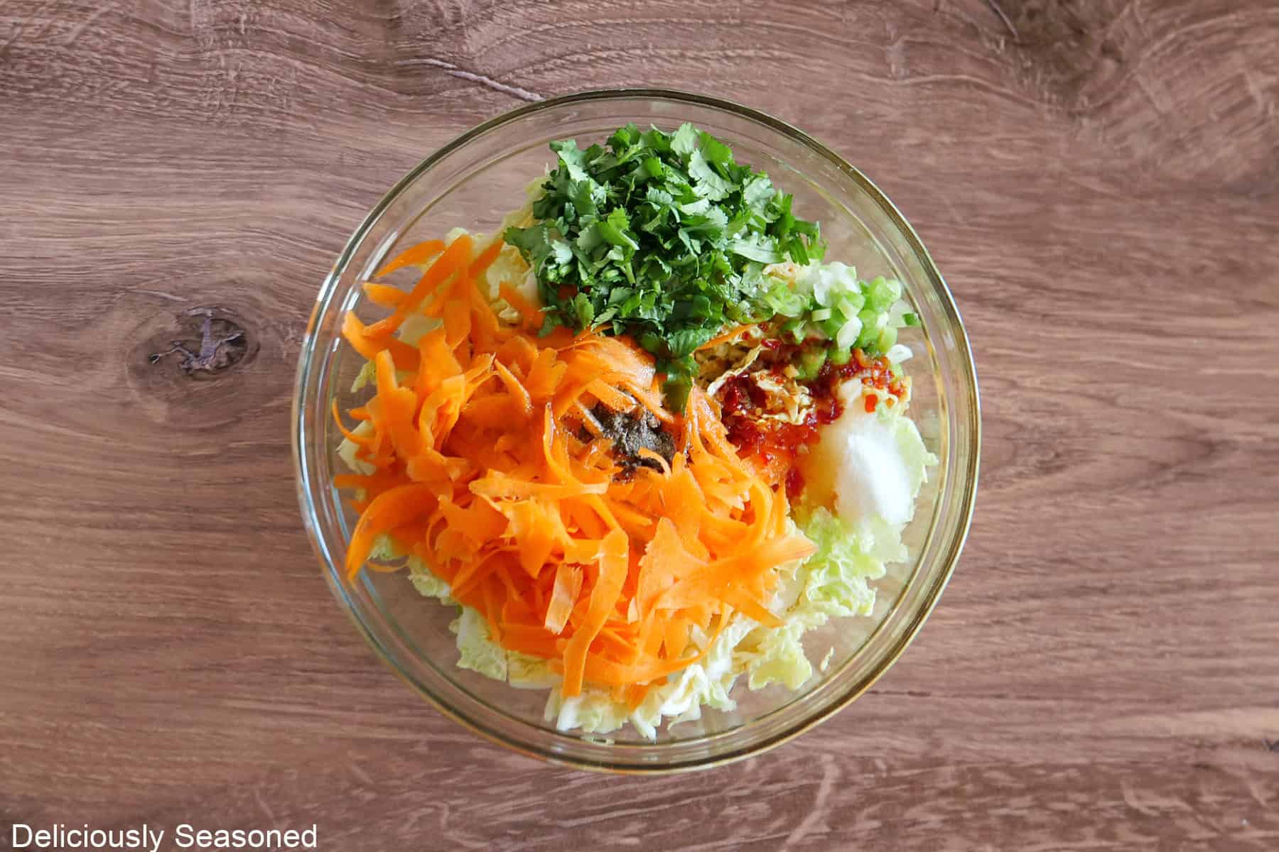 A glass bowl placed on a wood surface with the slaw ingredients placed in it.