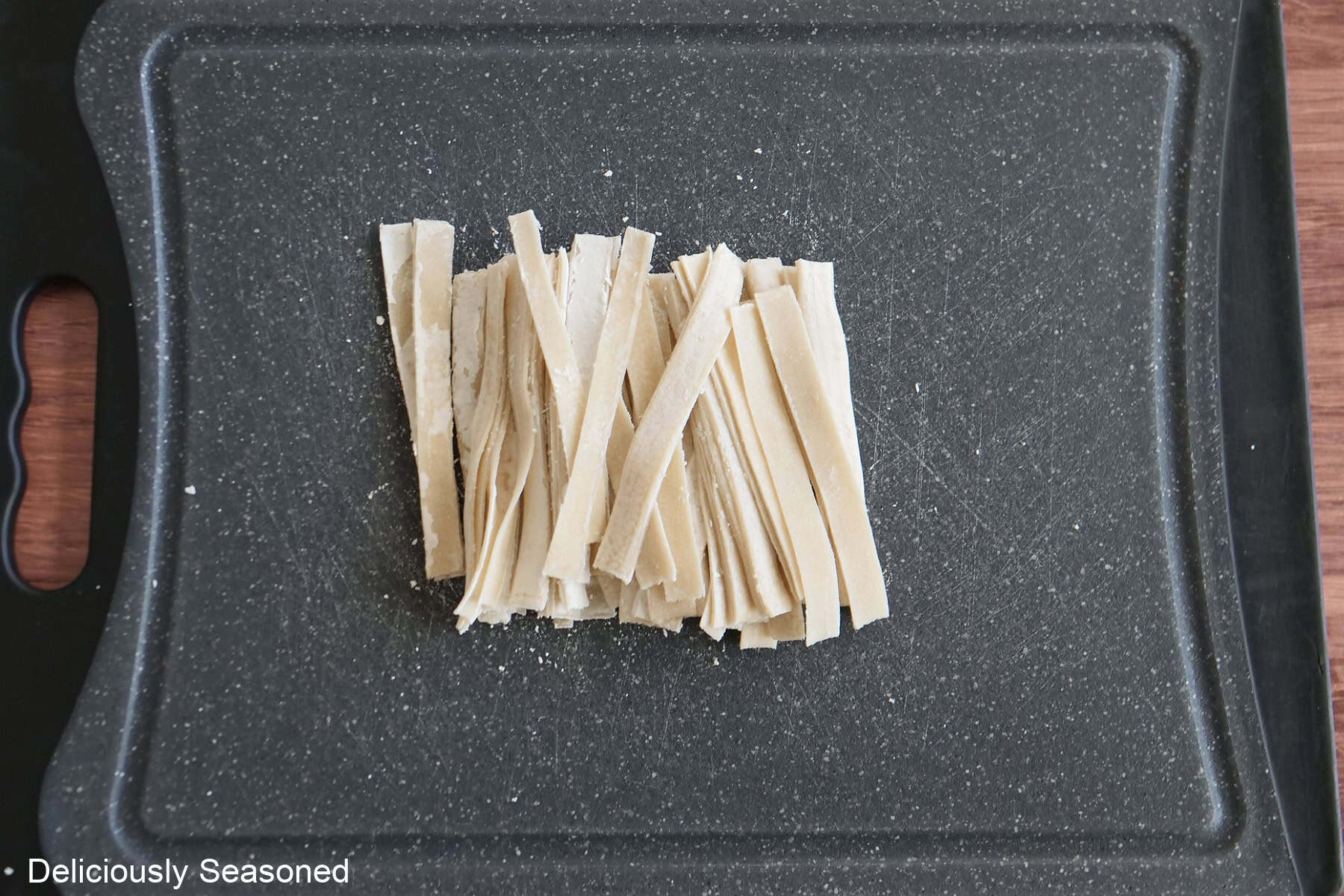 A black cutting board with wonton wrappers cut into thin strips.