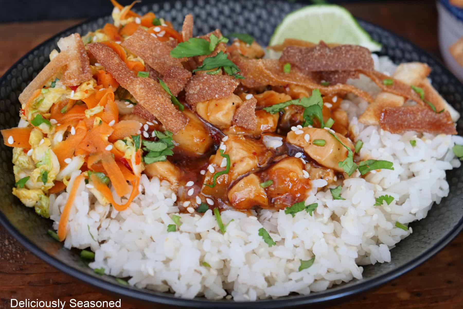 A black bowl filled with white rice and savory pieces of chicken with slaw and a chili sauce.