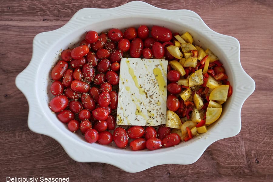 A white oval baking dish with grape tomatoes, squash, a block of feta cheese, and seasonings and olive oil.