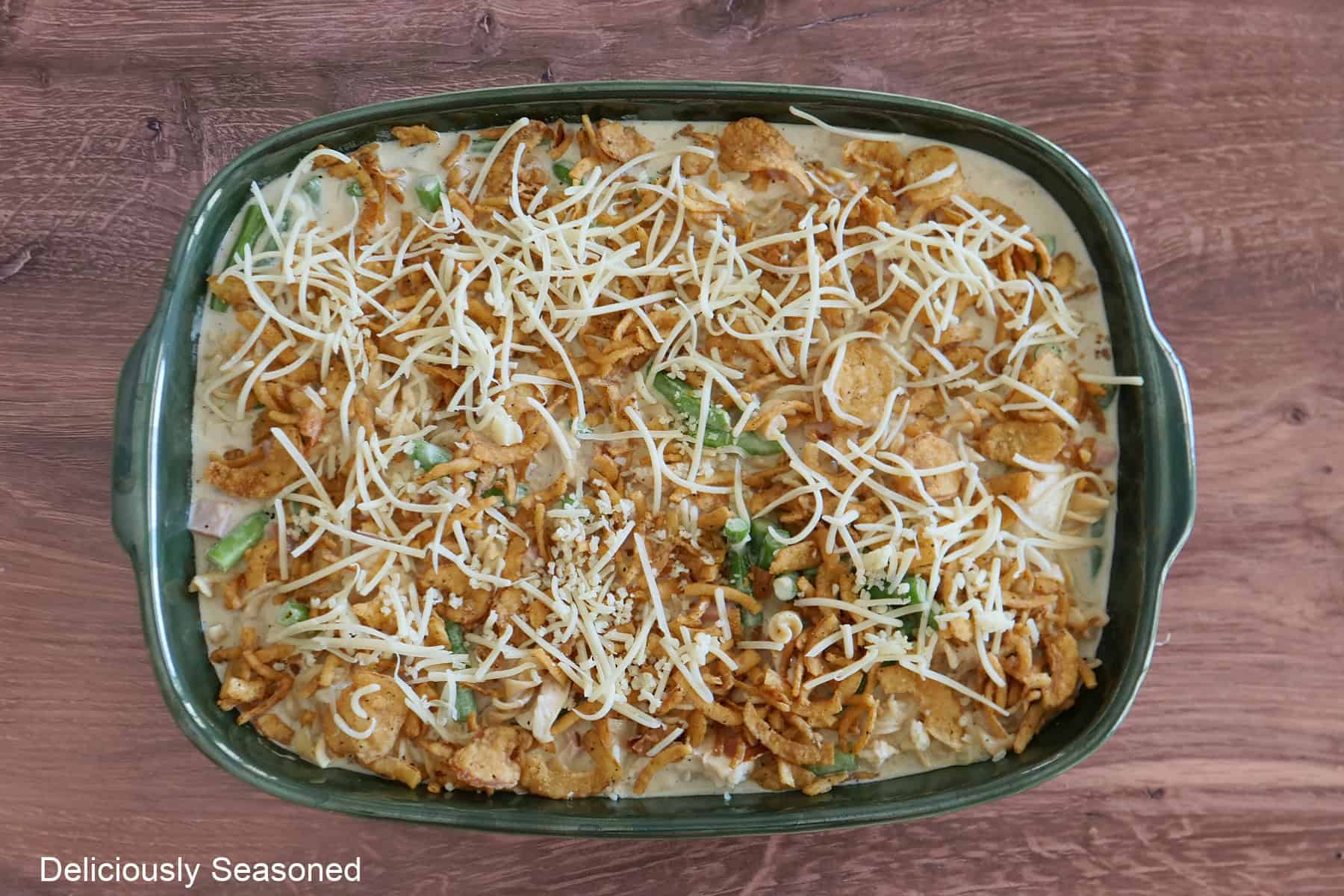 A wood surface with a large green baking dish filled with French onion chicken green bean casserole before going into the oven.