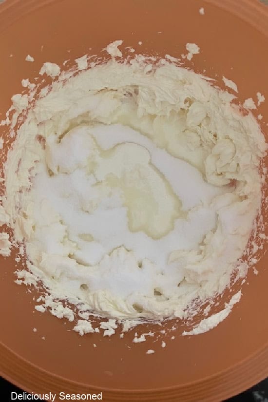 An orange mixing bowl with cream cheese that has been beat, with heavy cream and sugar added to the bowl.