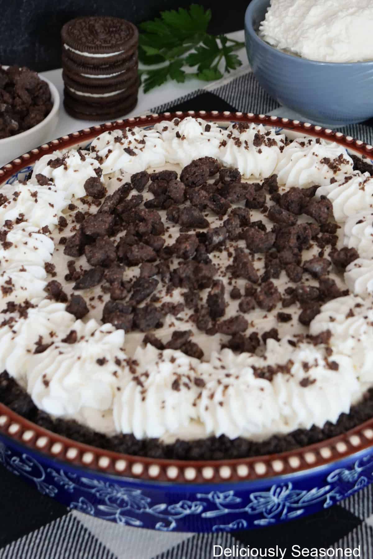 A blue floral pie dish with an Oreo cookie cream cheese pie in it topped with whipped cream and chocolate crunch topping.