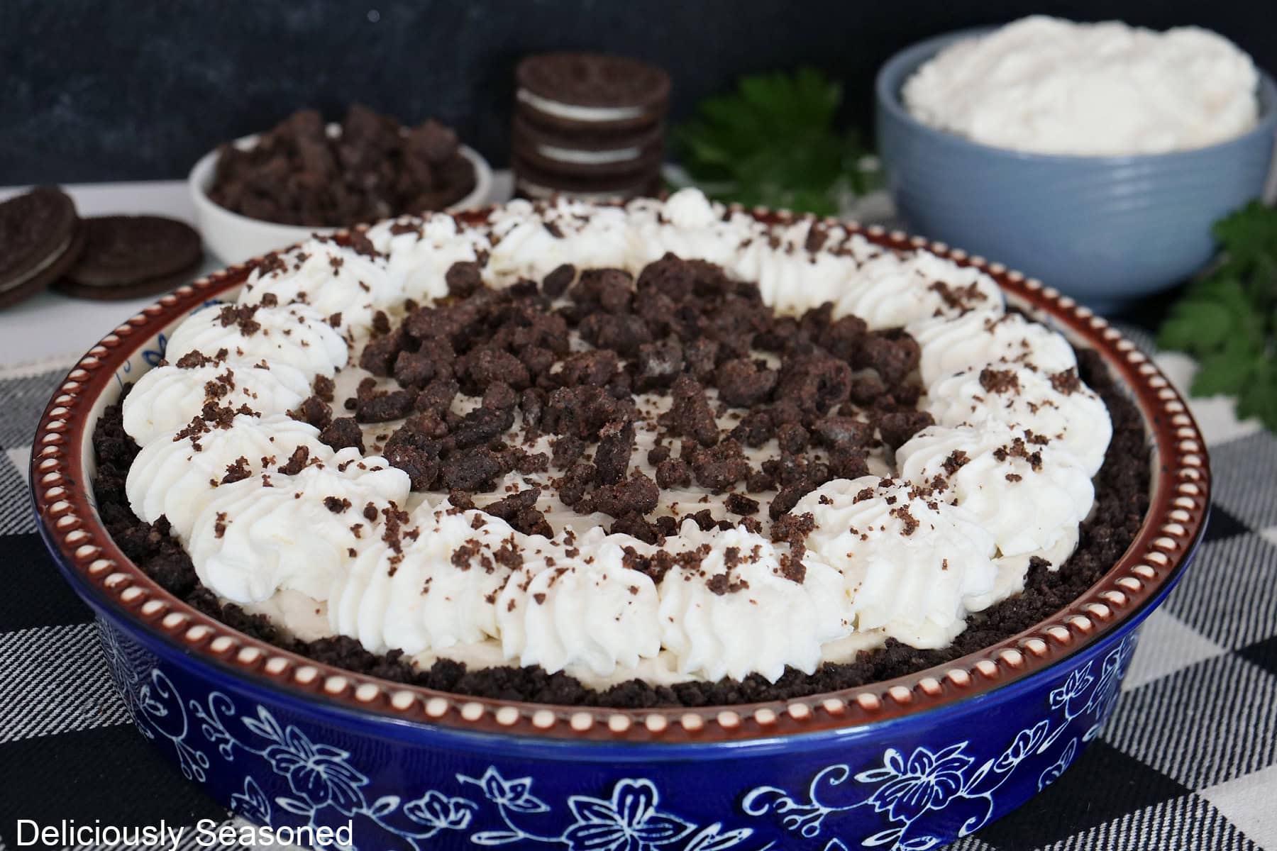 A white surface with a black and white checker placemat on it with a blue floral pie dish filled with an Oreo crust, a no-bake pie filling, homemade whipped cream, and chocolate crunch topping.