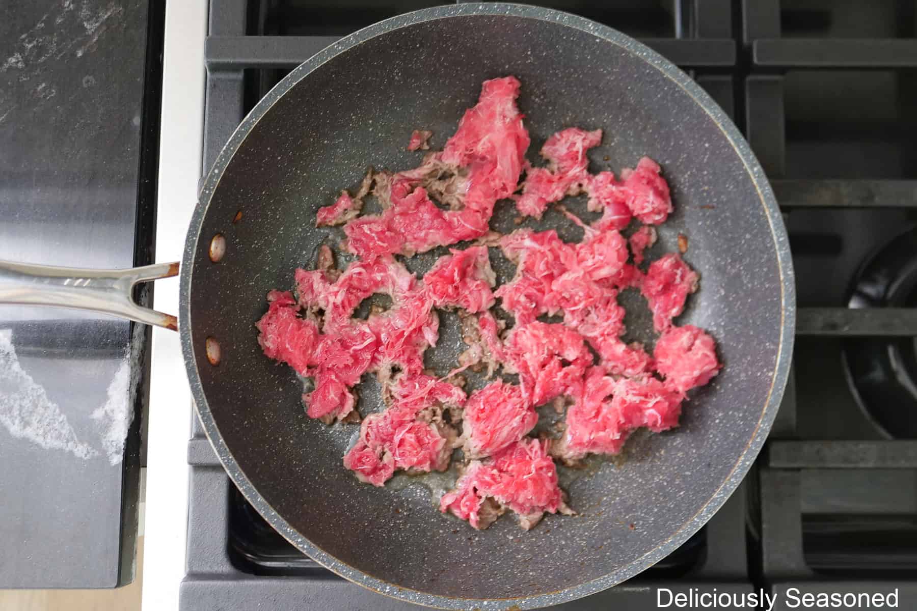 A frying pan with shaved beef in it cooking on the stove.