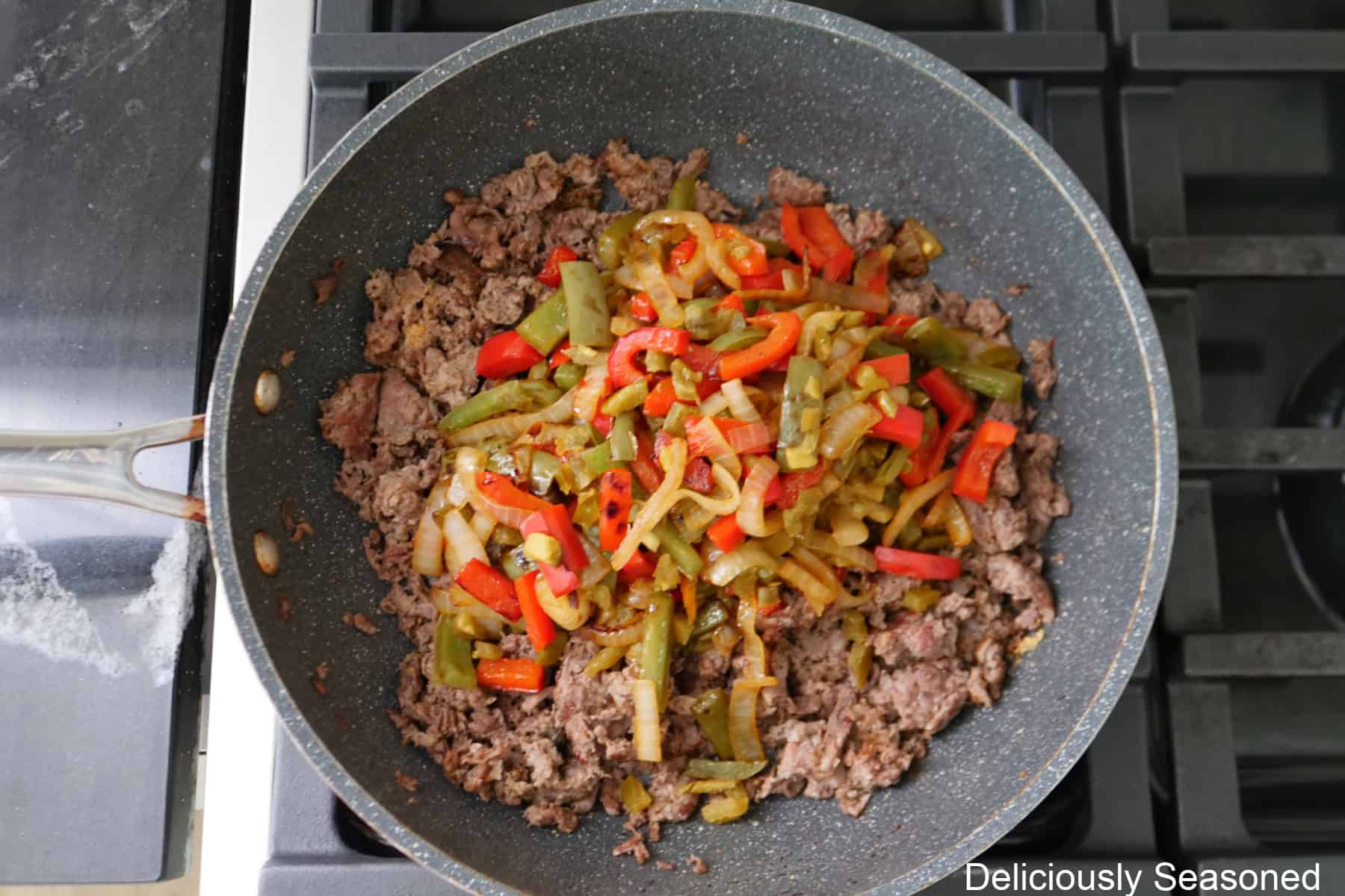 A frying pan with cooked beef, peppers and onions, and diced jalapenos in it.