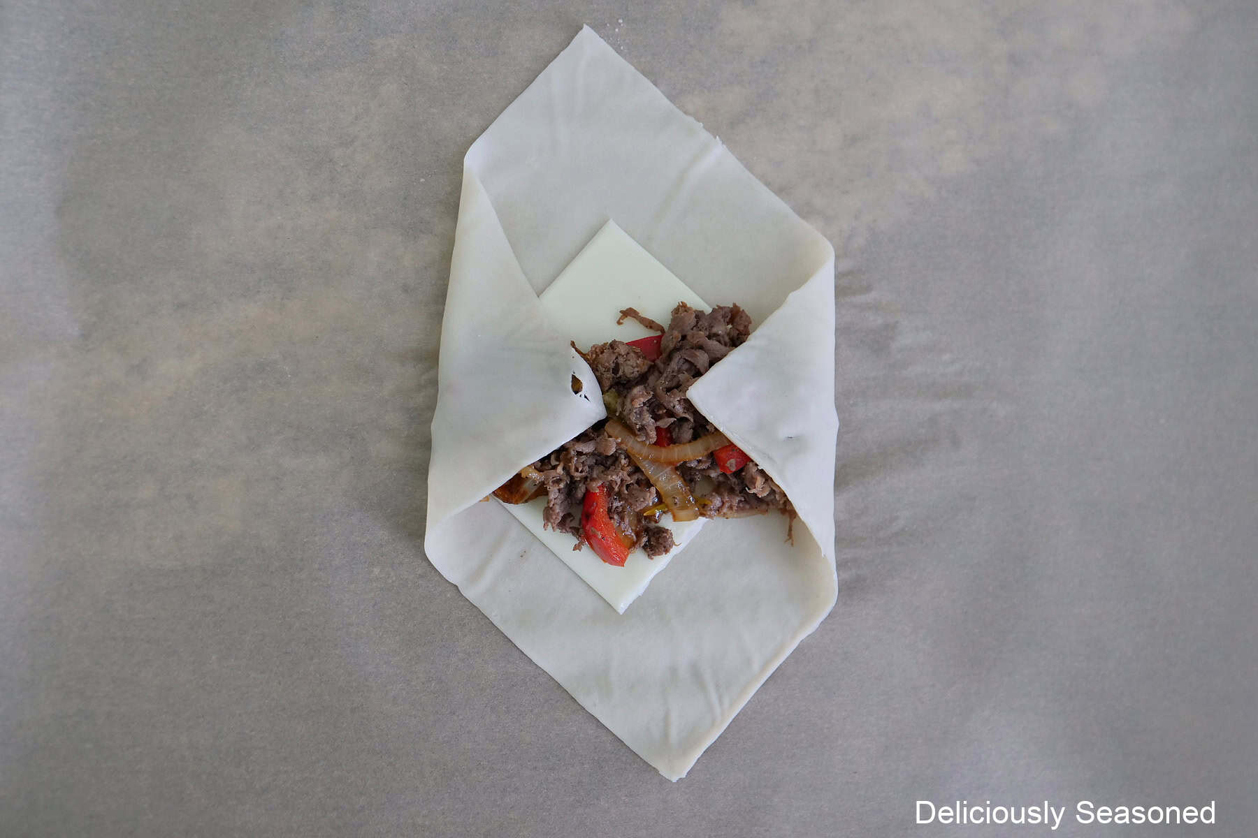 An egg roll wrapped on a piece of parchment paper with cheese and beef mixture on it and two corners folded over the meat mixture.