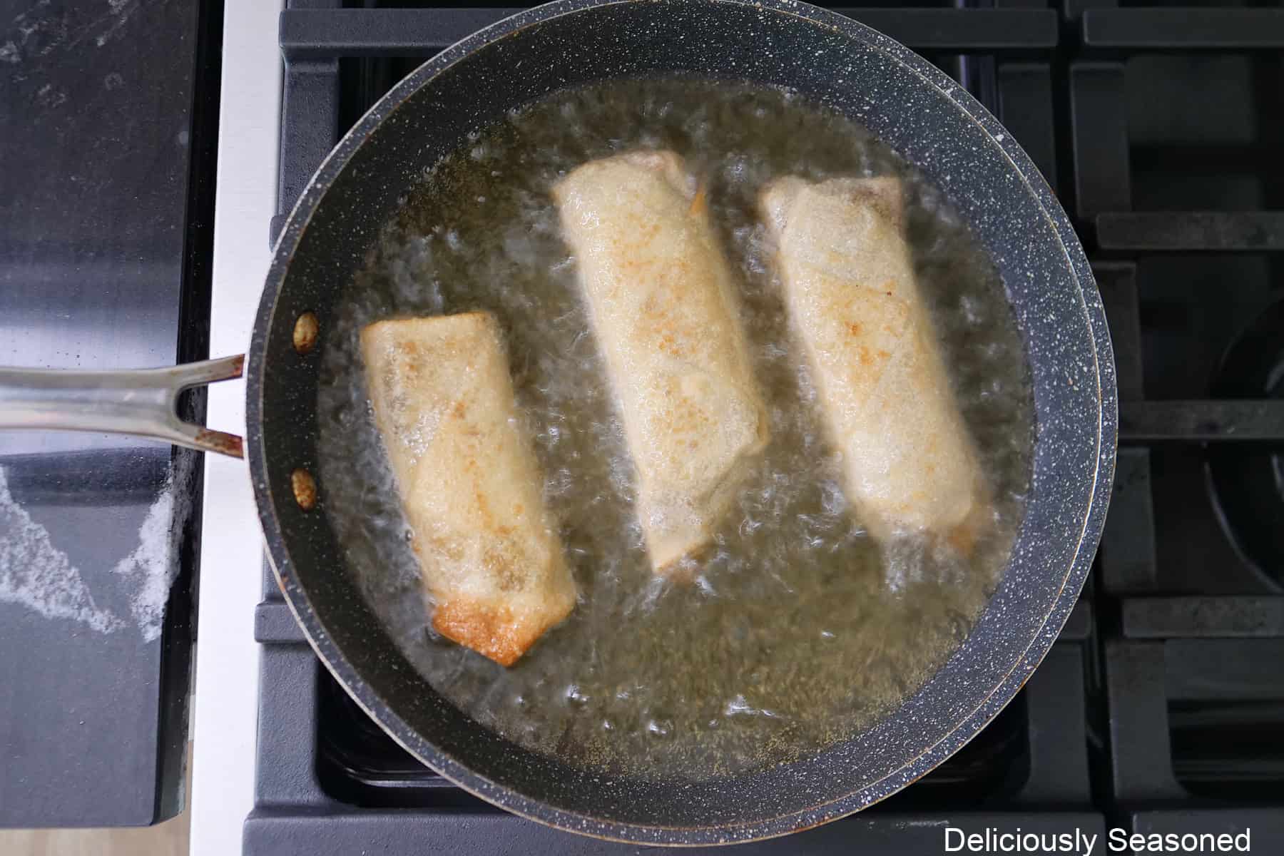 A frying pan with three egg rolls in it frying in oil.