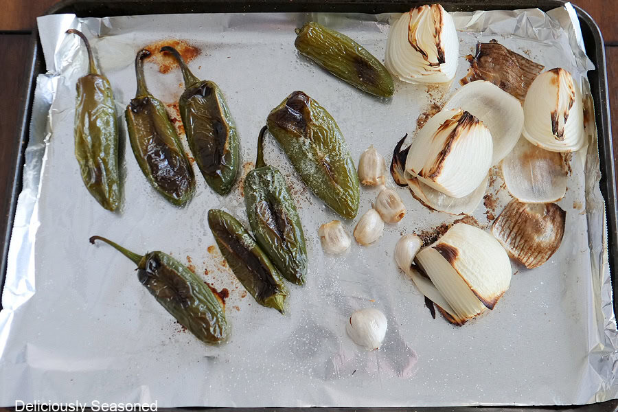A foil lined baking sheet with roasted jalapenos, onions, and garlic cloves on it.