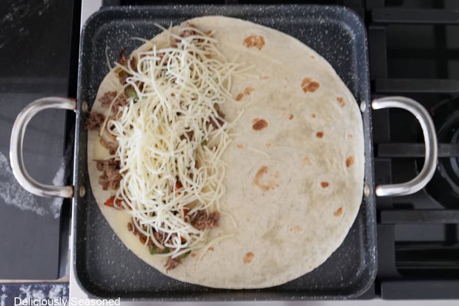 A flour tortilla in a pan with cheesesteak beef and shredded cheese on one side.