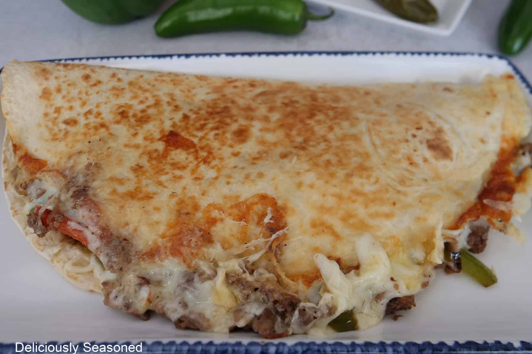 A golden fried Philly cheesesteak quesadilla on a white plate with blue trim before being cut into three pieces.