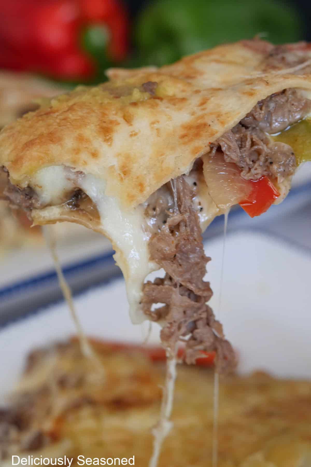 A slice of Philly cheesesteak quesadilla held up off the plate showing the gooey, melty cheese and cheesesteak hanging out of the tortilla.