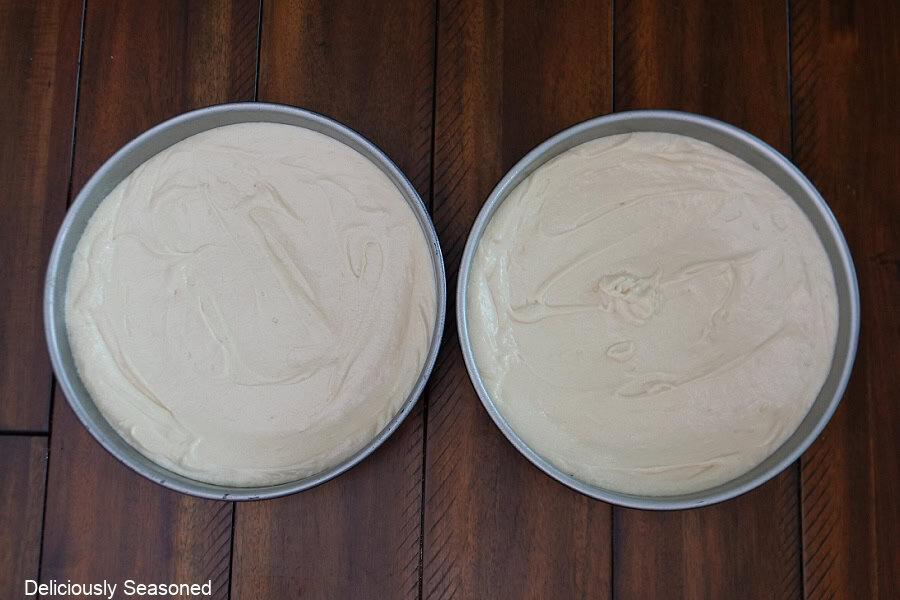 Two round cake pans with cake batter in them both placed on a wood surface.