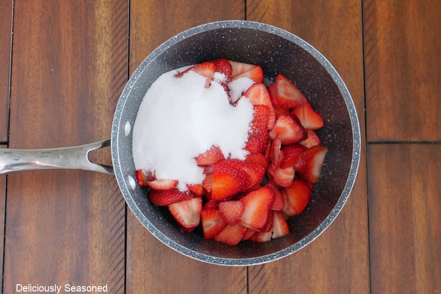 A saucepan with freshly sliced strawberries and sugar in it, place on a wood surface.