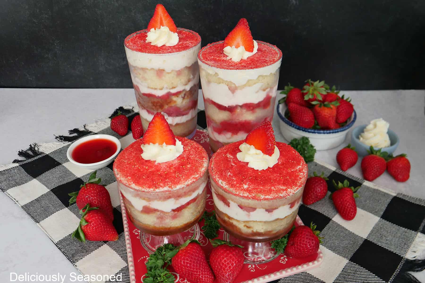 Two small and two tall strawberry tiramisu parfaits on a black and white checkered placemat with a red ceramic base and fresh strawberries placed around.