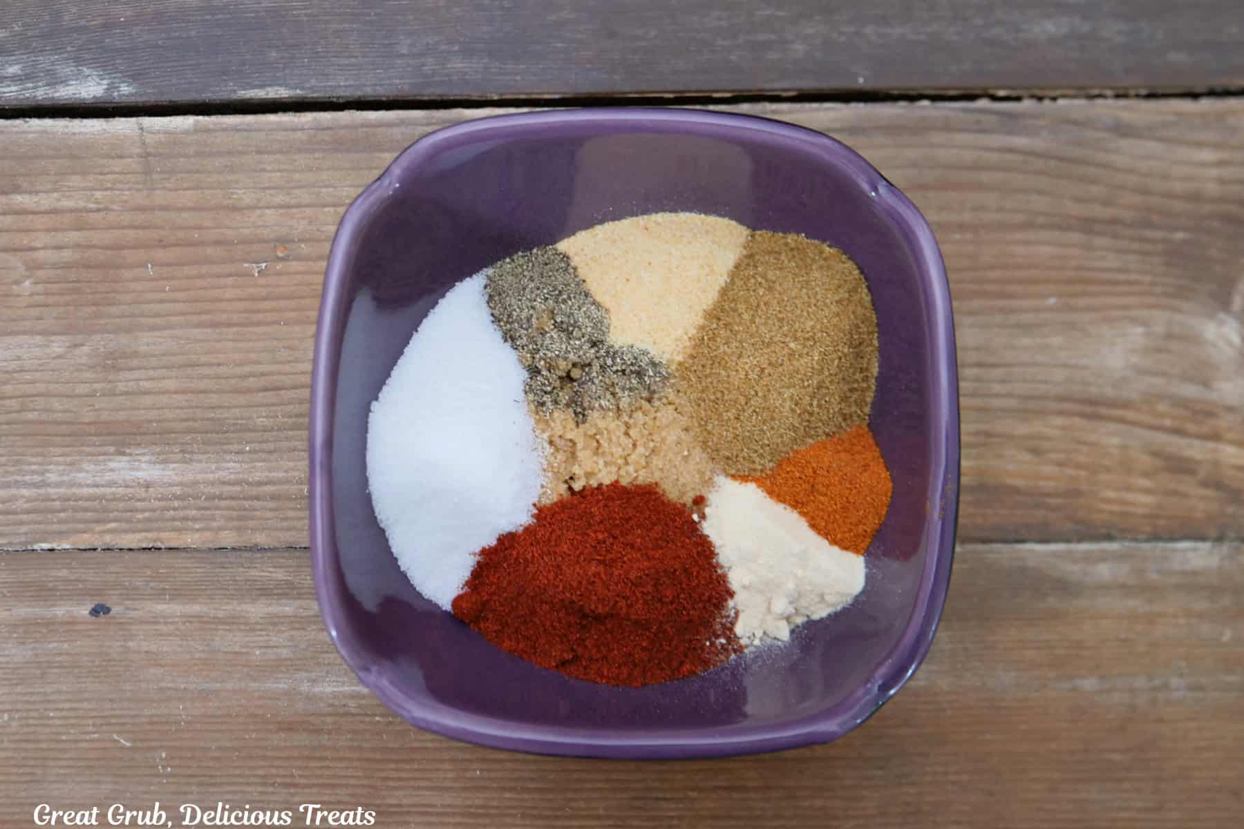 A wood surface with a small purple bowl filled with all the different seasonings that gets rubbed on the ribs.
