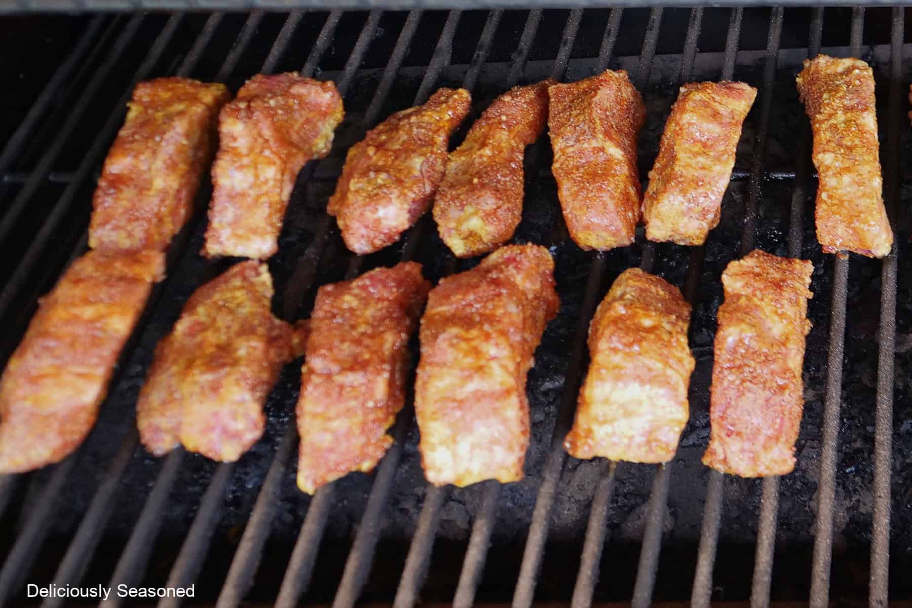 Thirteen ribs on the grill grates of a smoker being smoked.