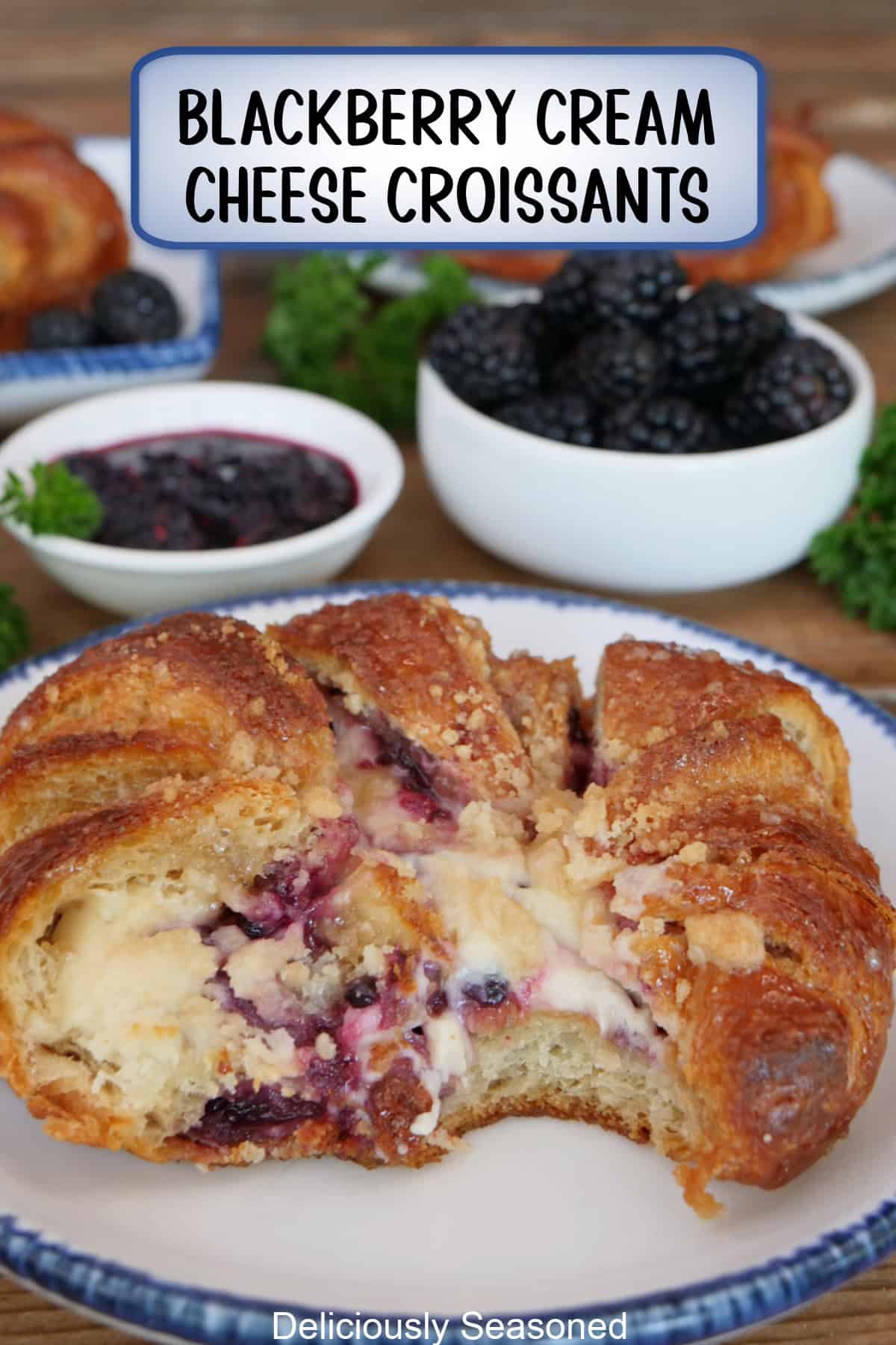 A white plate with blue trim with a round blackberry cream cheese croissant with two white small bowls in the background with blackberries, and a title overlay at the top.