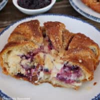 A round white plate with blue trim with a blackberry cream cheese croissant on it with a few bites taken out of it.