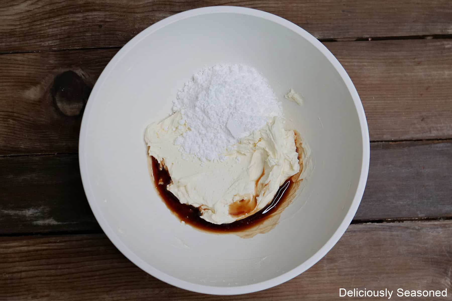 A wood surface with a white mixing bowl placed on it with the cream cheese mixture ingredients in it before being mixed together.