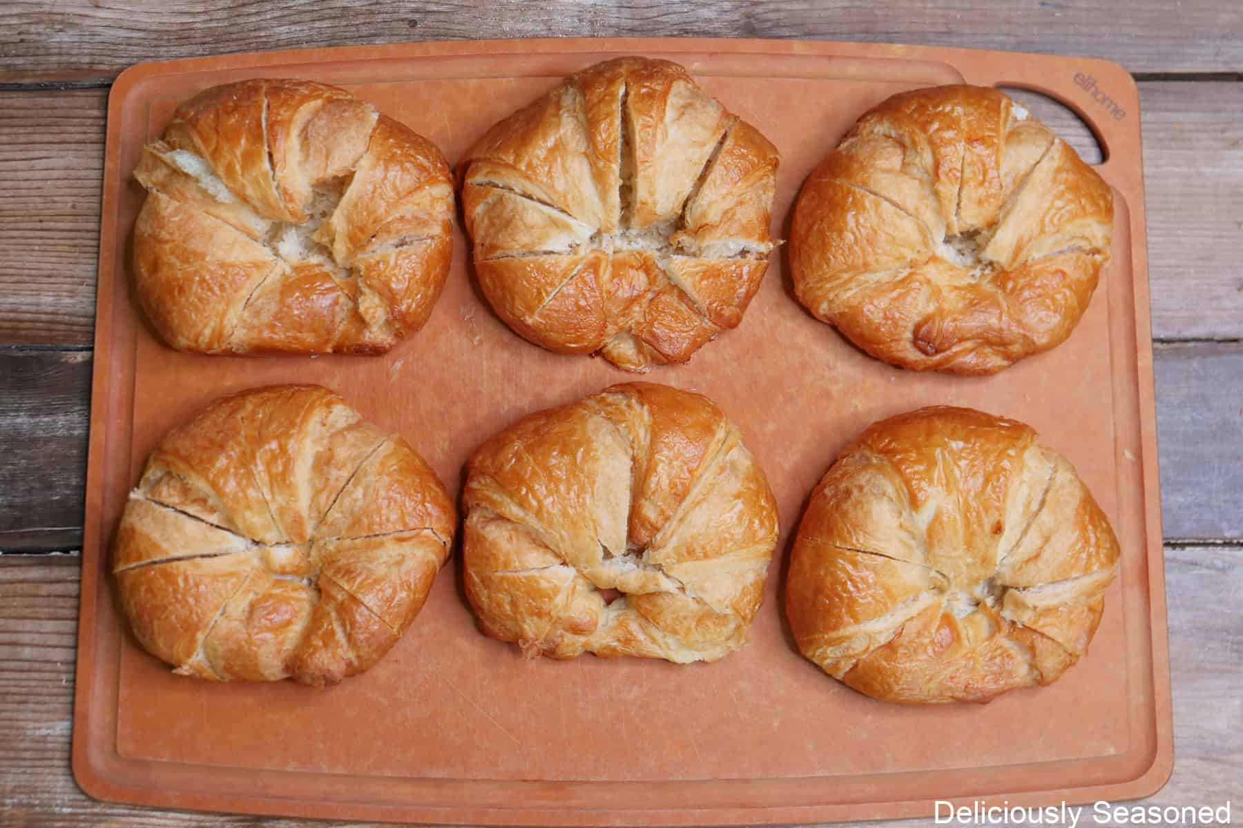 A cutting board with six round store-bought croissants on it with cuts in the tops of each croissant.