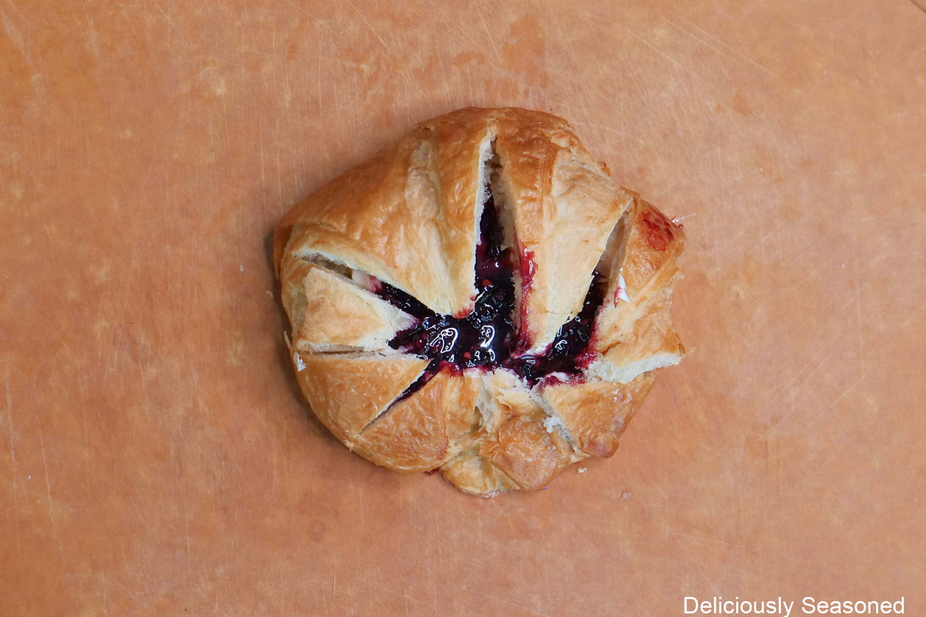 A cutting board with one round croissant on it filled with a sweet cream cheese mixture and a blackberry filling.