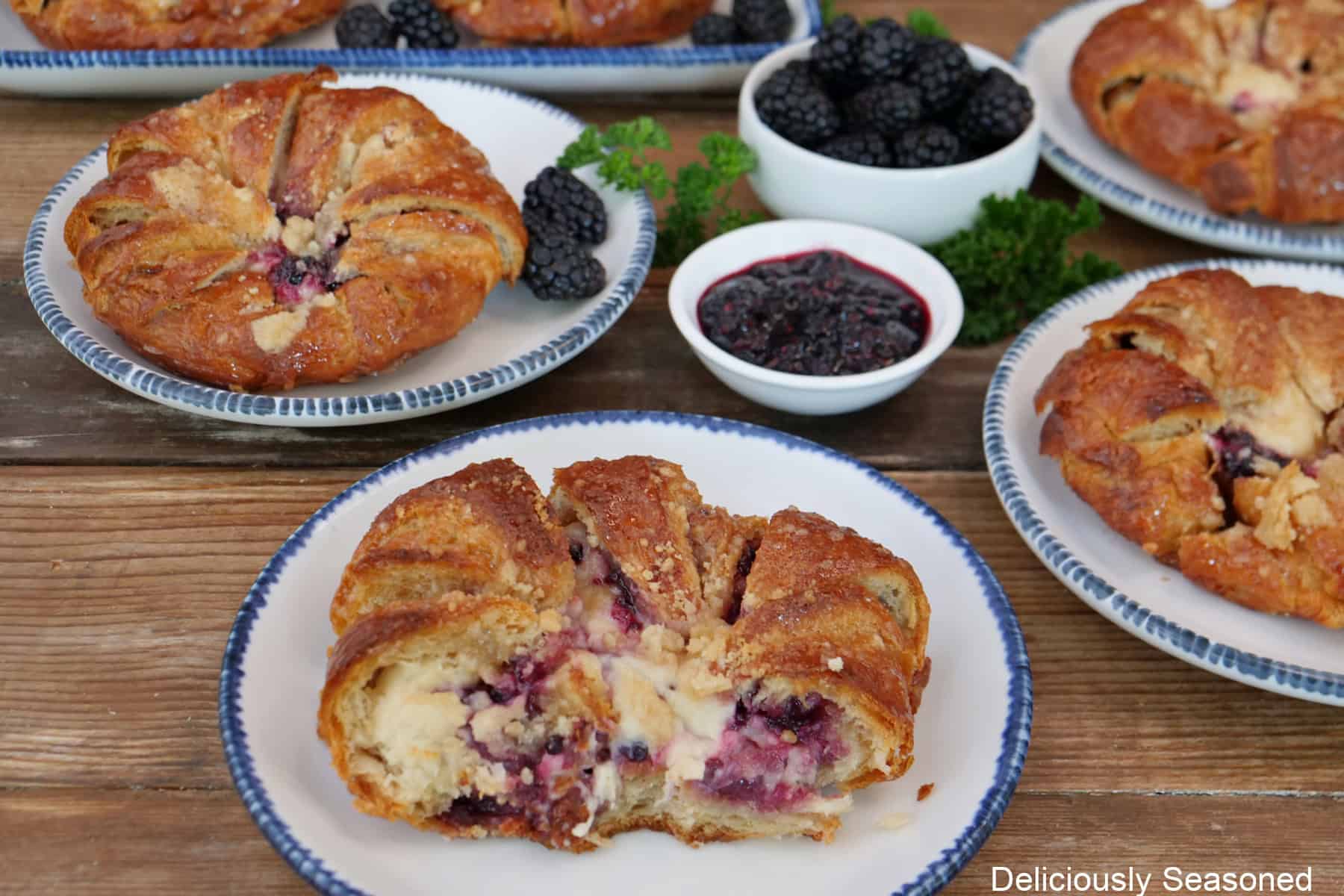 A wood surface with a few white plates with blue trim with blackberry cream cheese croissants on them with two white bowls with blackberries and blackberry filling in them.