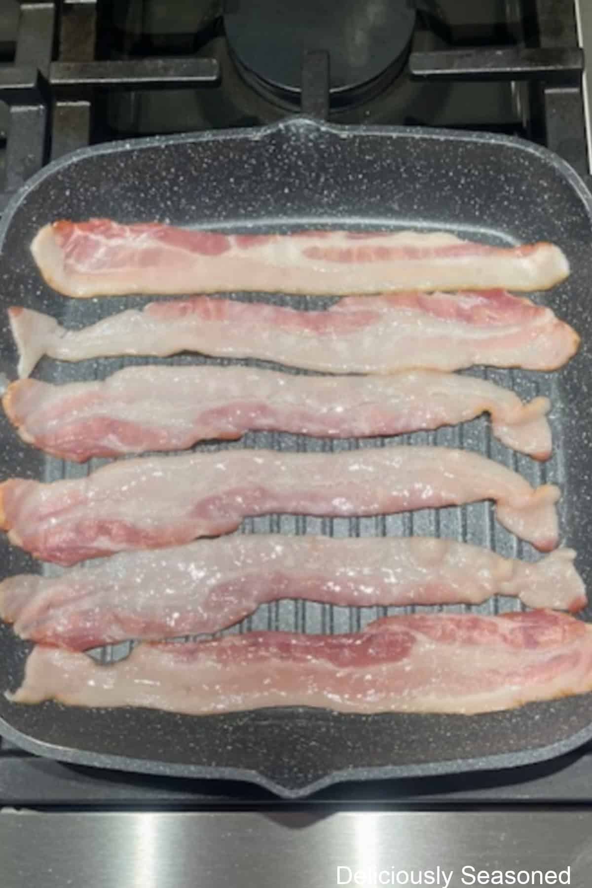 A shallow pan on the stove with six strips of bacon in it cooking.