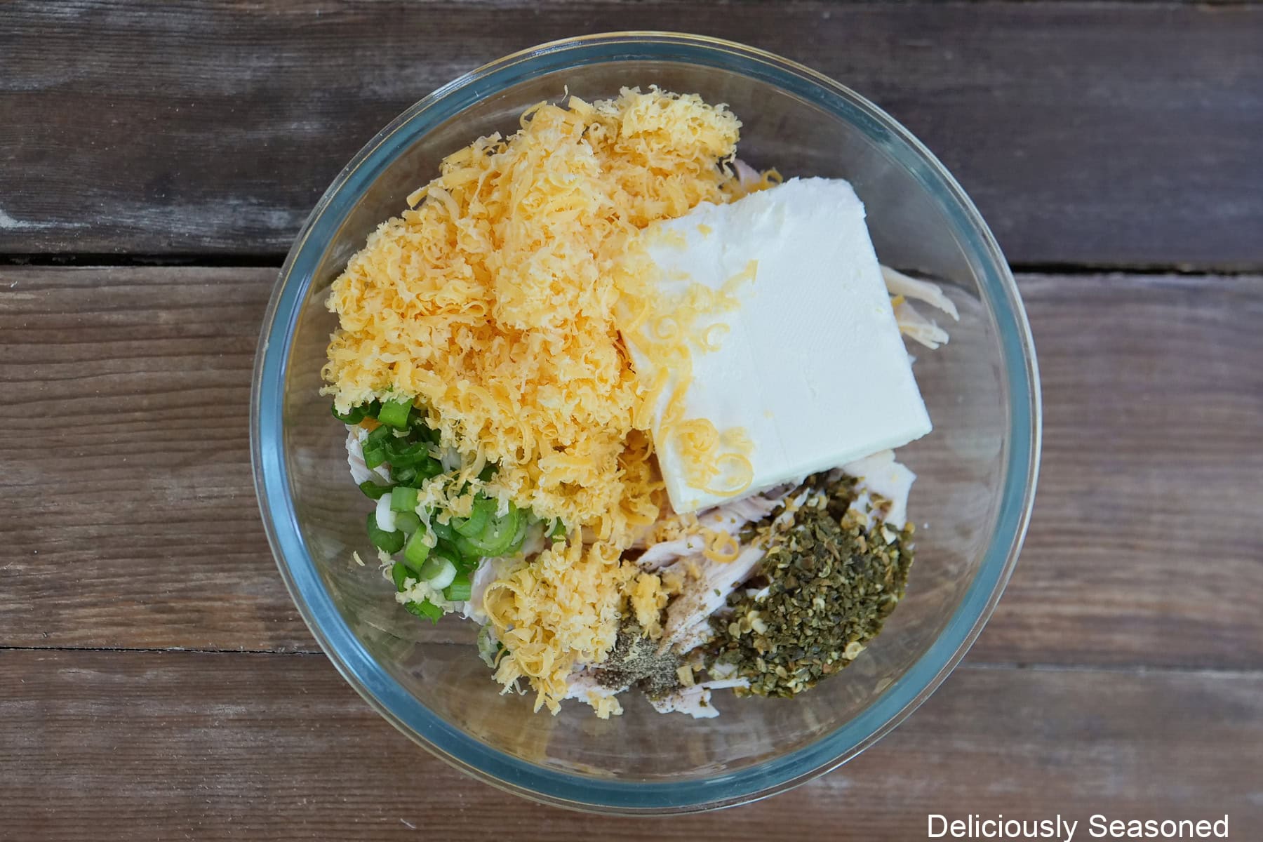 A wood surface with a glass bowl filled with shredded rotisserie chicken, cheddar cheese, cream cheese, sliced green onions, and jalapeno seasoning, before being mixed together.