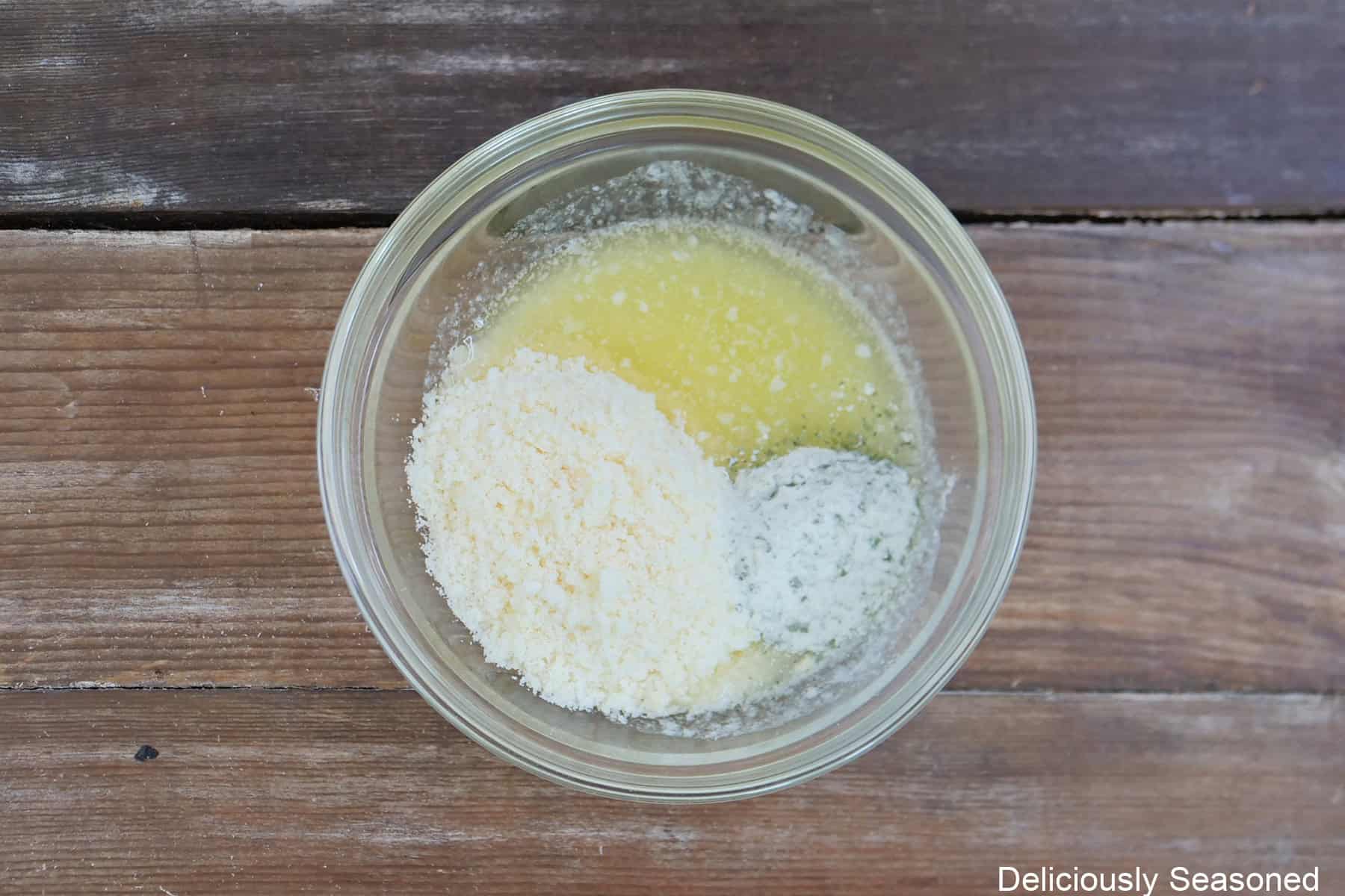 A wood surface with a glass bowl with melted butter, ranch dressing, and Parmesan cheese in it.
