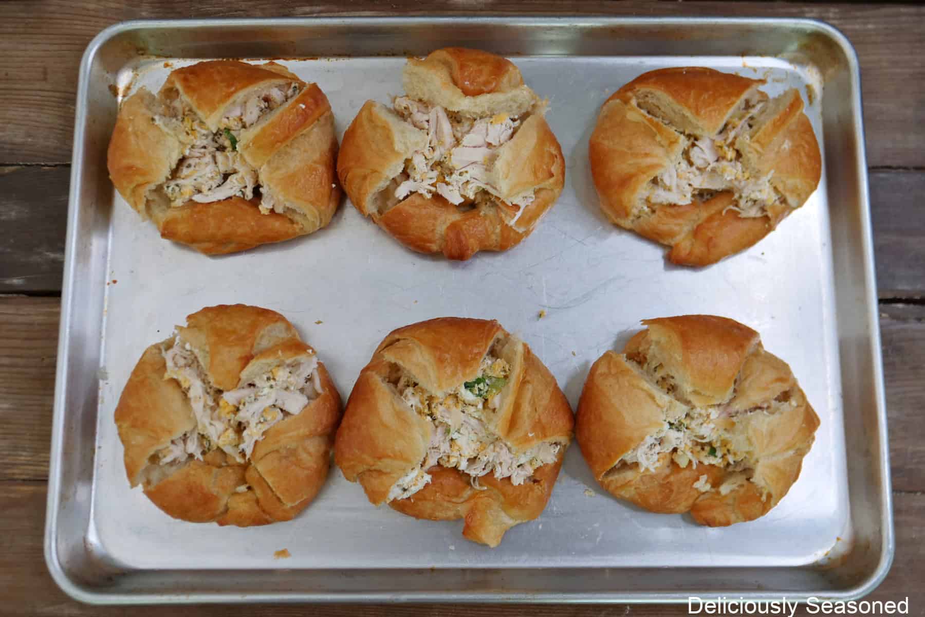 A silver baking sheet with six croissants filled with a rotisserie chicken mixture before being baked in the oven.
