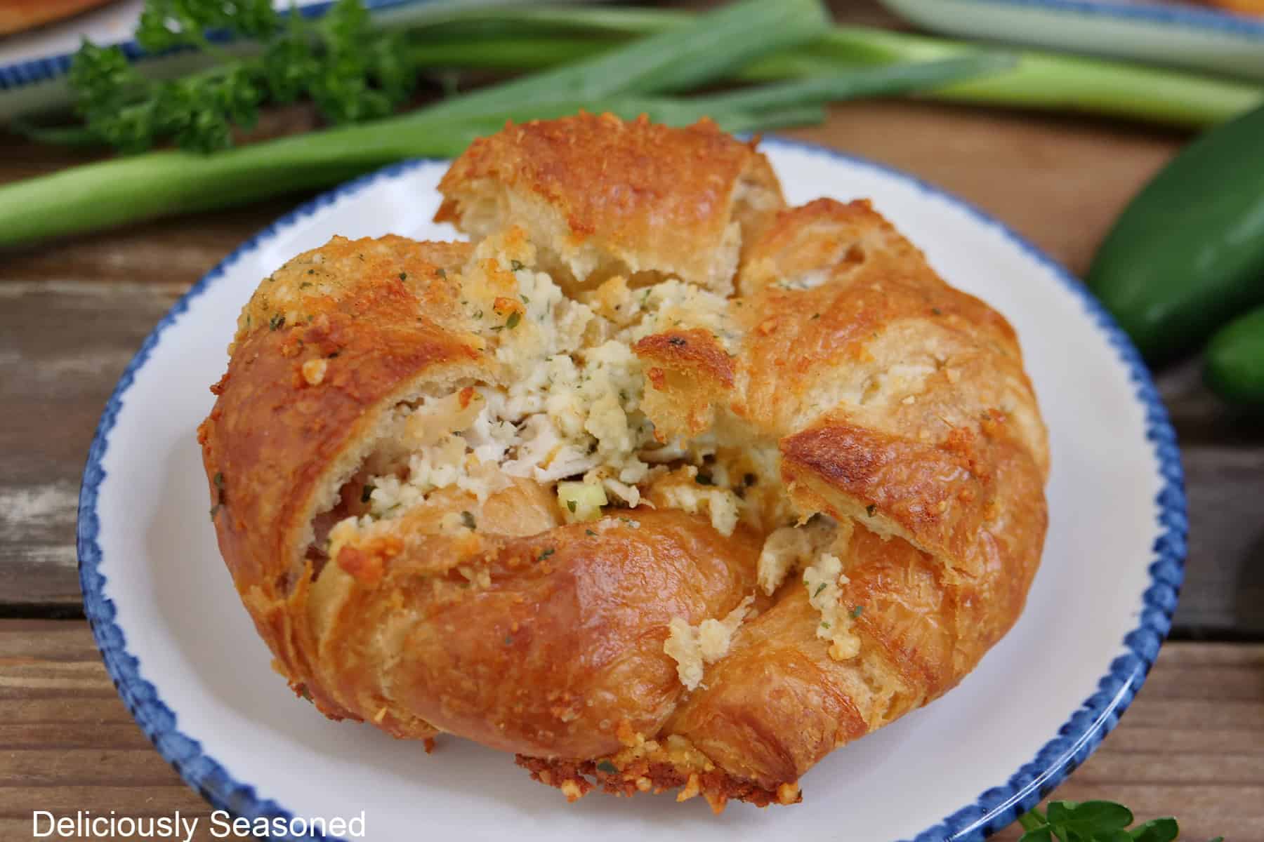 A white plate with blue trim with a baked chicken ranch crusted croissant on it with green onions in the background.