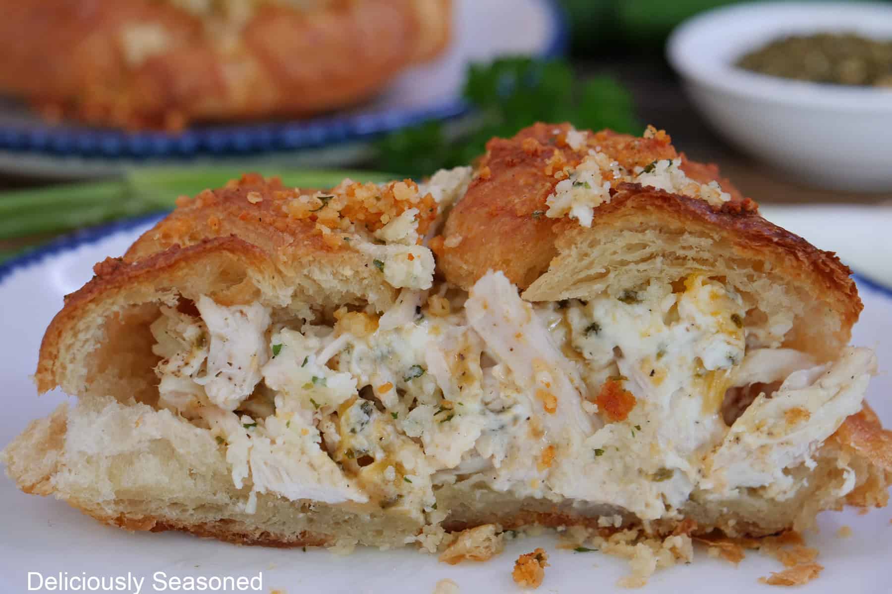 A white plate with blue trim with a chicken ranch crusted croissant on it that has been cut in half to show the savory inside ingredients.