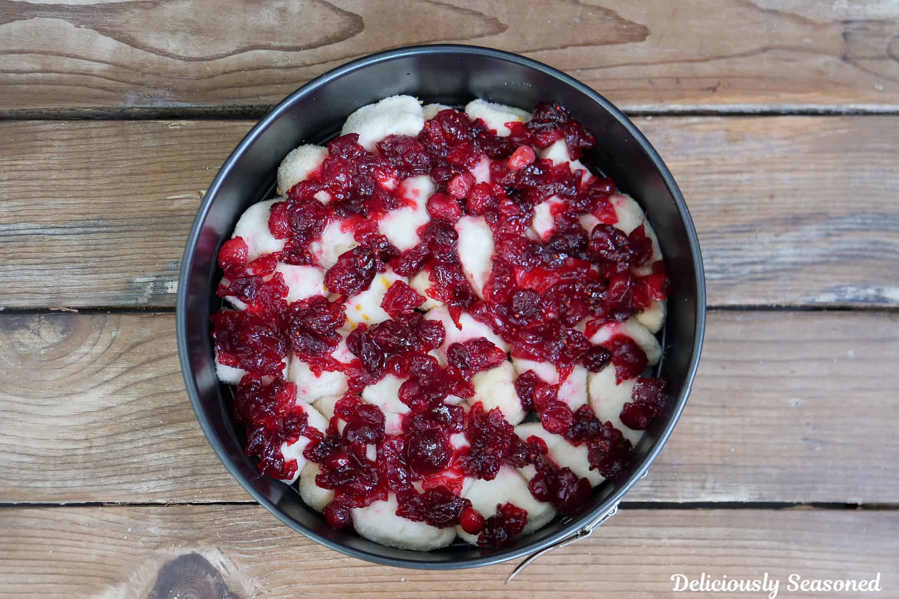 A rustic wood surface with a springform pan filled with coated biscuit pieces, and topped with cranberry orange mixture.
