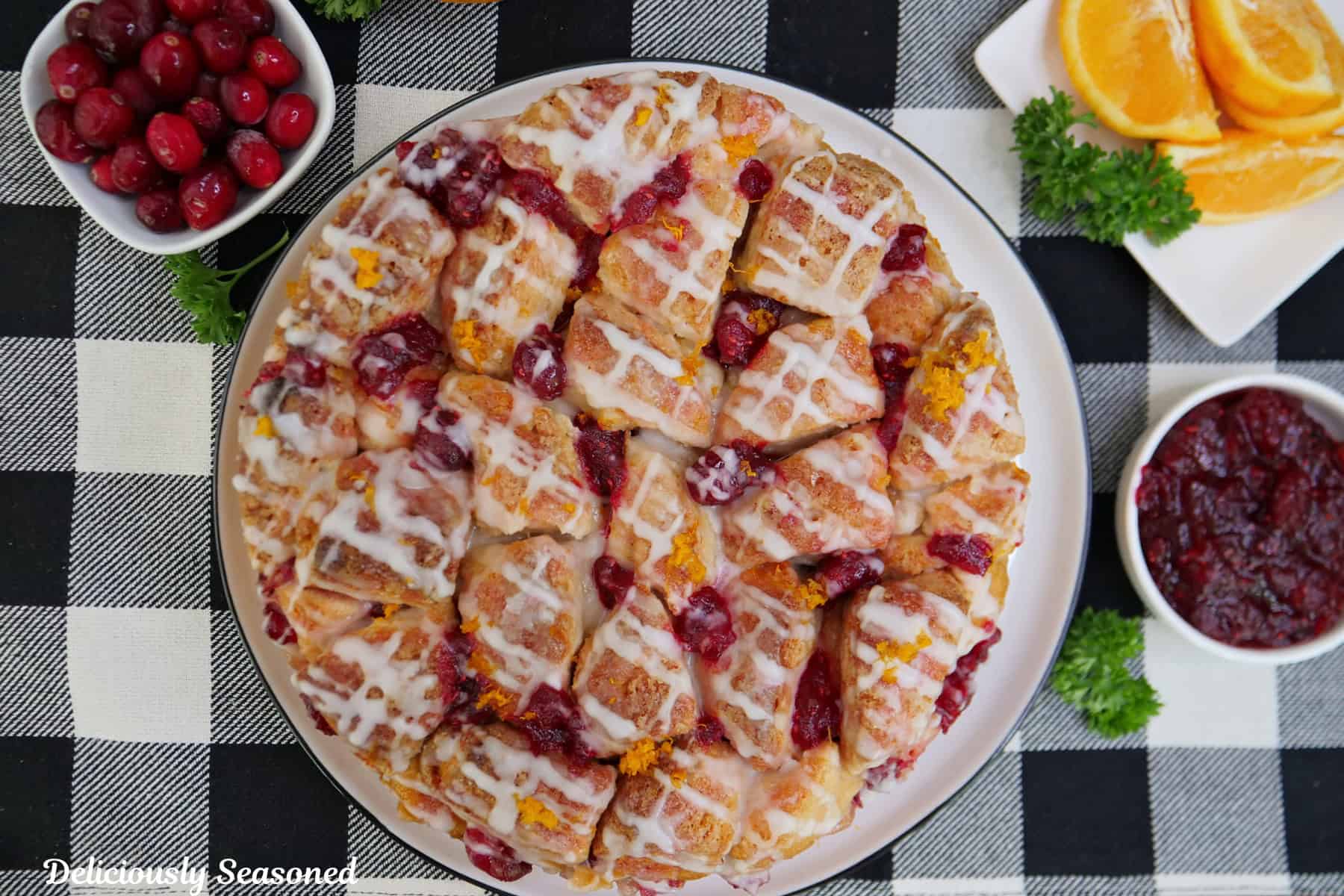 A black and white checkered placemat with a cranberry orange monkey bread placed on a white plate with cranberries and cranberry filling in white small bowls beside it, with orange slices on a white plate.