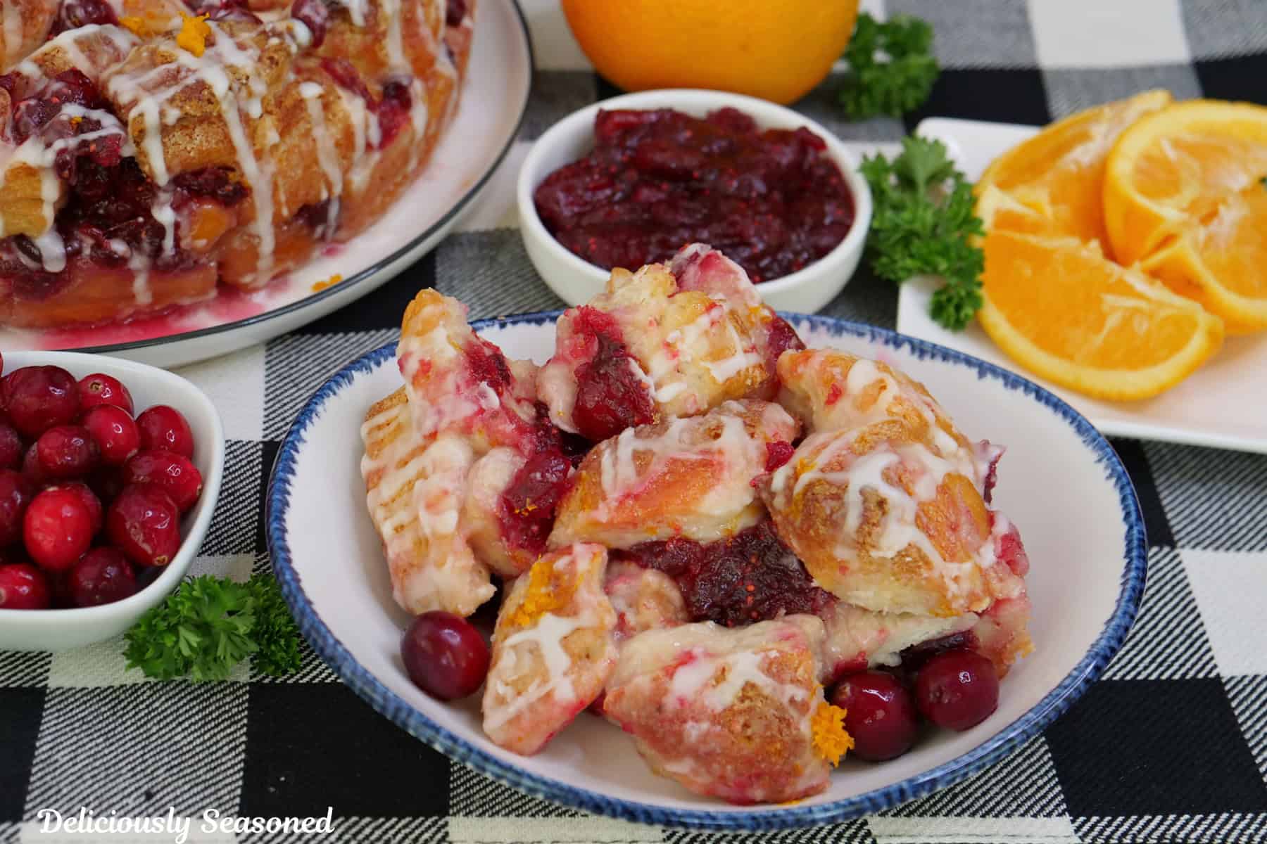 A black and white checkered placemat with a white bowl with blue trim filled with a serving of cranberry orange monkey bread in it, with orange slices, cranberries, and cranberry filling in the background.