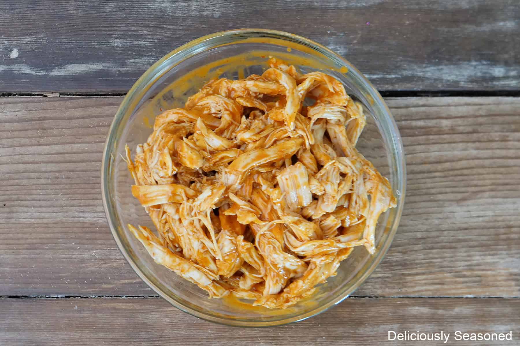 A glass bowl filled with shredded rotisserie chicken with buffalo sauce mixed in to it placed on a wood surface.