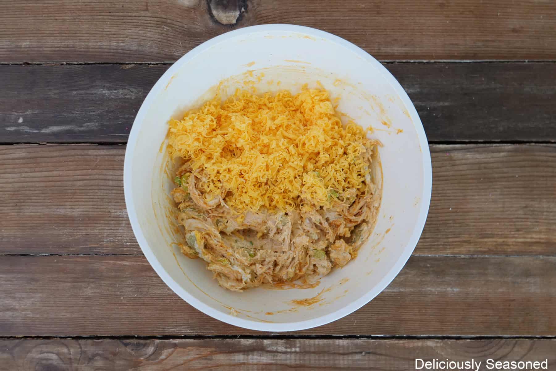 A rustic wood surface with a white bowl filled with a shredded chicken mixture with shredded cheese on top.