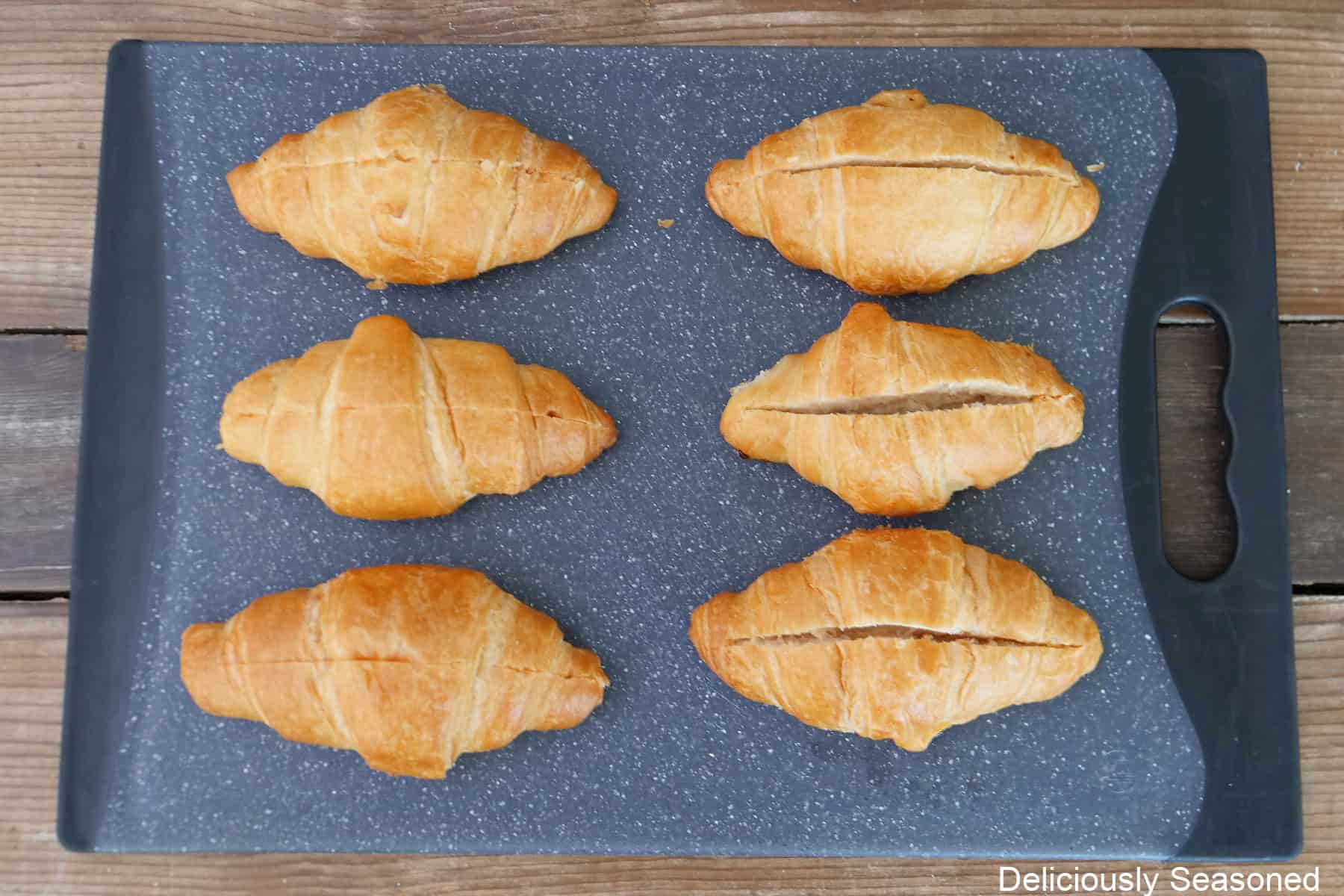 A black cutting board with six mini croissants placed on it that are made with a slice in the top of each one.