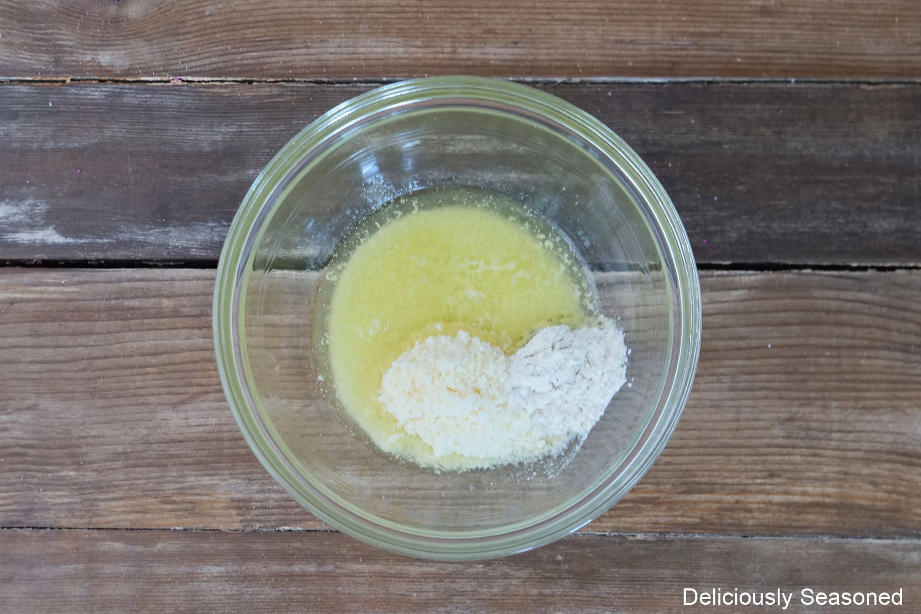 A glass bowl placed on a rustic wood surface with melted butter, Parmesan cheese, and ranch seasoning mix in it.
