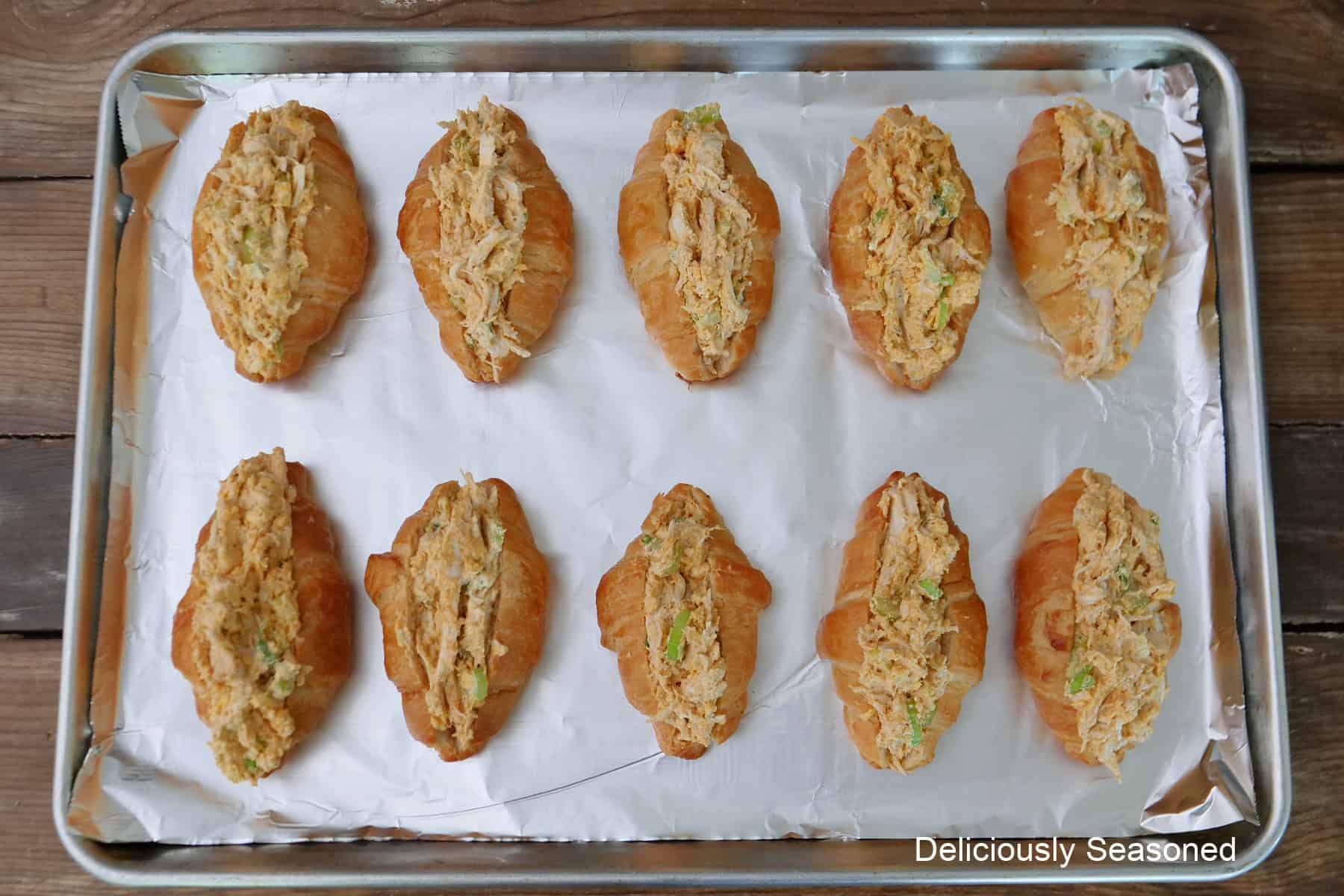 A wood surface with a foil lined baking sheet with ten min croissants filled with buffalo chicken mixture.