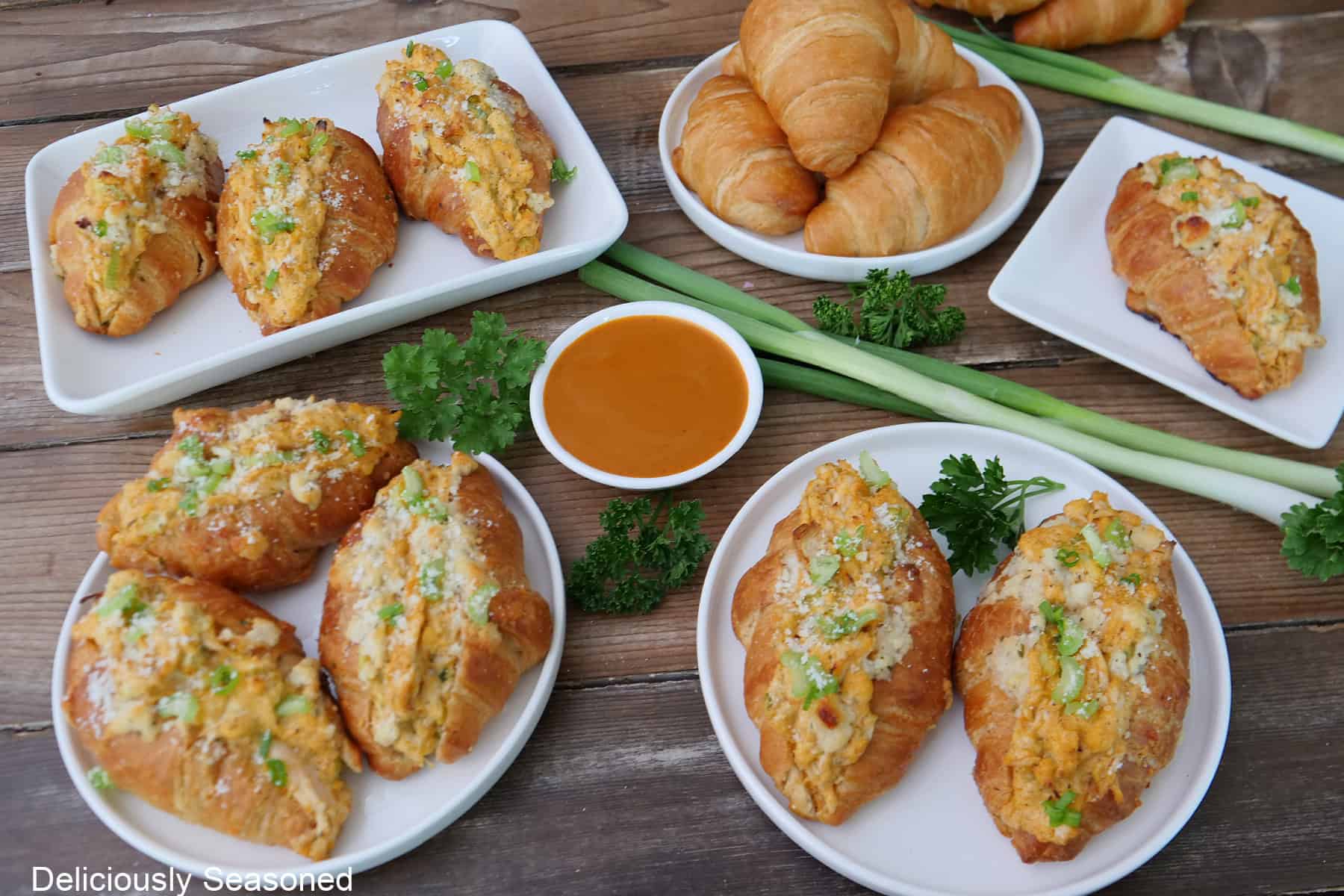 A rustic wood surface with mini buffalo chicken croissants placed on white plates with a small white bowl filled with buffalo wing sauce and green onions and parsley in the background.